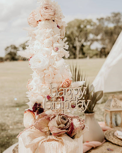 Elegant wooden "happy birthday" topper adorned with pastel flowers, set outdoors against a soft, natural backdrop.