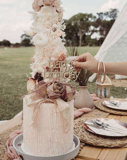 A hand places a wooden "happy birthday" topper on a beautifully decorated pastel cake adorned with flowers.