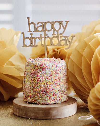 A small, colorful cake with a "happy birthday" topper, surrounded by soft, yellow decorations on a wooden base.