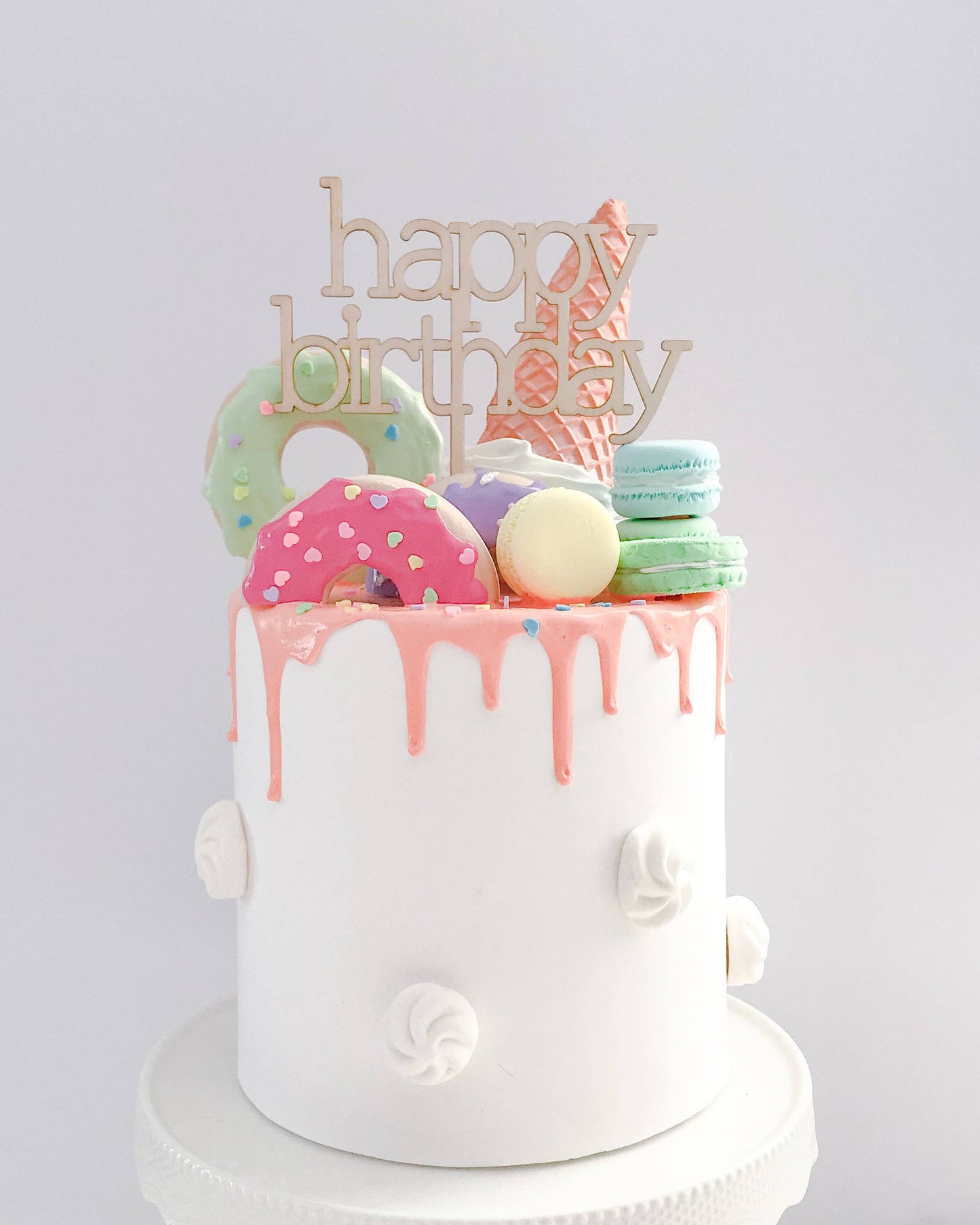 A white cake topped with colorful sweets and a "happy birthday" wooden topper, featuring pink icing drips.