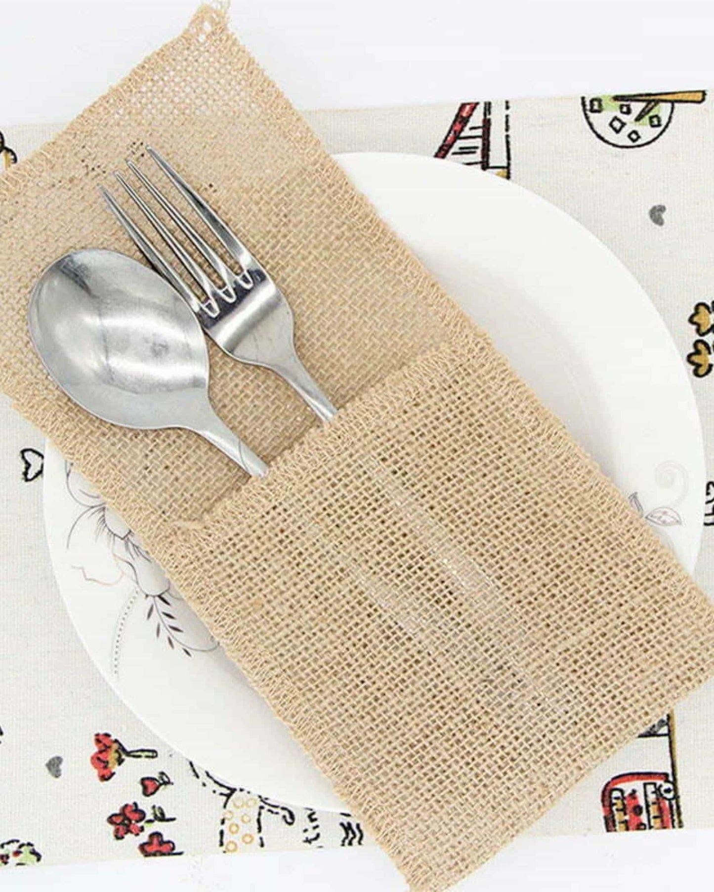 A burlap pouch holds a spoon and fork atop a decorative plate on a patterned tablecloth.