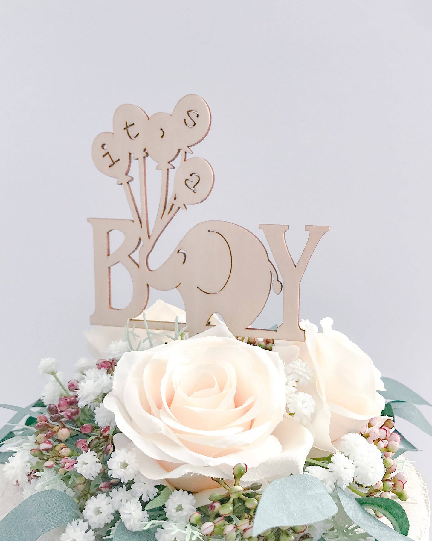 A wooden elephant cake topper with balloons, surrounded by soft pastel roses and greenery, celebrating a baby boy.