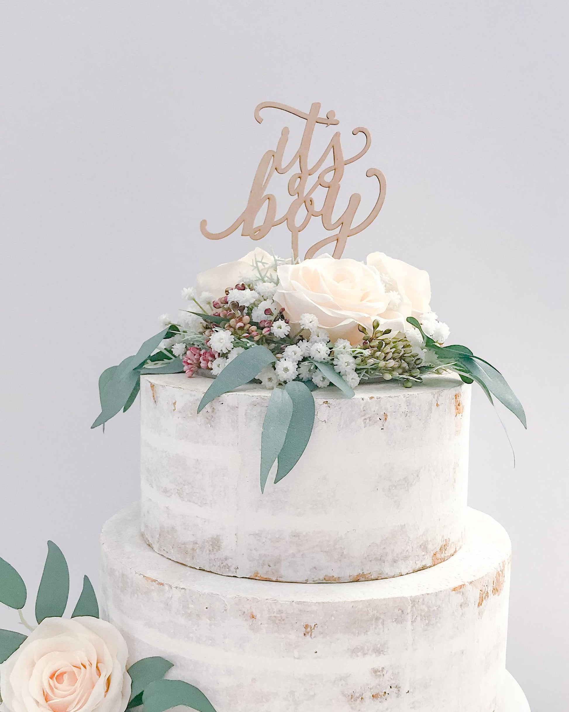 A wooden topper reads "It's a boy" atop a layered cake adorned with pale roses and delicate greenery.