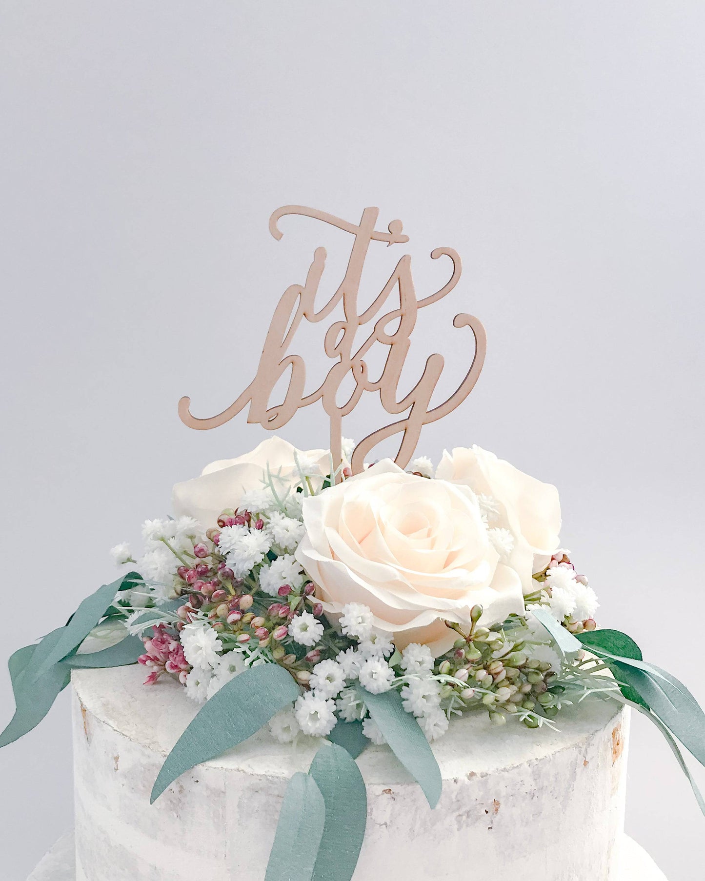 Wooden topper with "it's a boy" phrase atop a cake adorned with soft roses and delicate greenery.