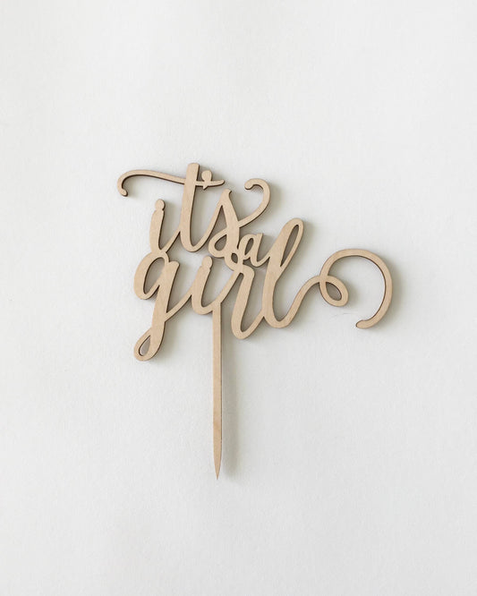 Wooden cake topper with elegant cursive text reading "it's a girl" on a light, neutral background.