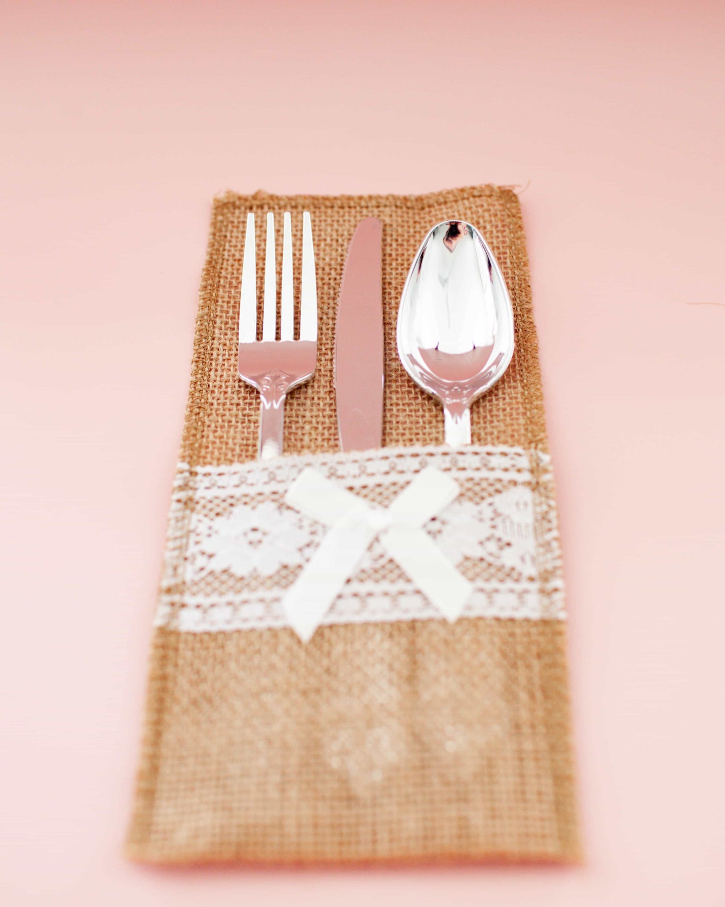 A burlap pouch holds a fork, knife, and spoon, adorned with lace and a delicate bow on a soft pink background.