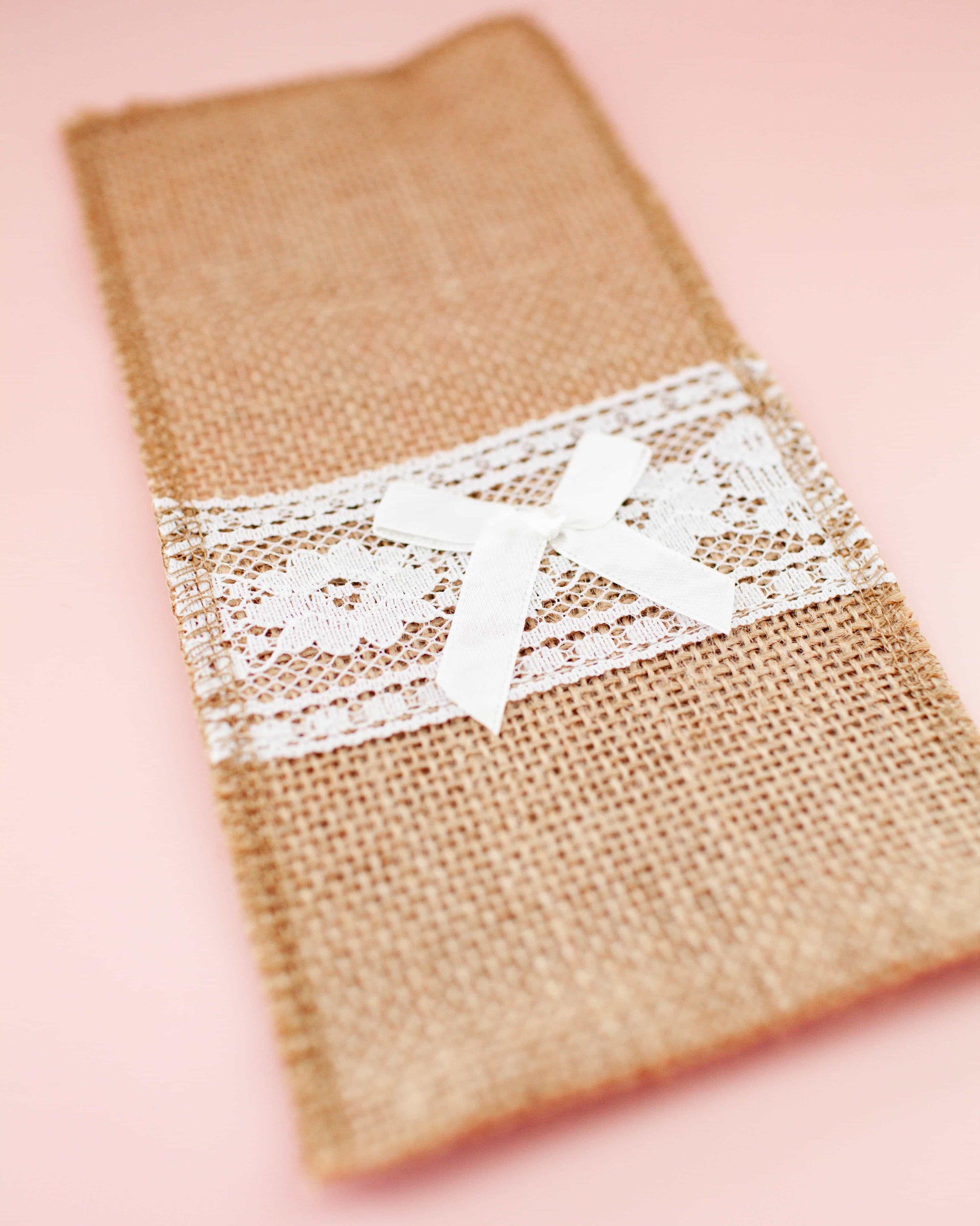 Linen cutlery holder in natural hessian with lace trim and a delicate white bow. Soft pink background enhances elegance.