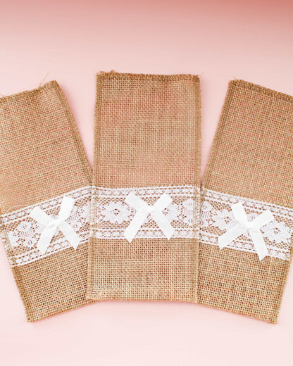 Three burlap cutlery wraps featuring delicate white lace and bows, set against a soft pink background.