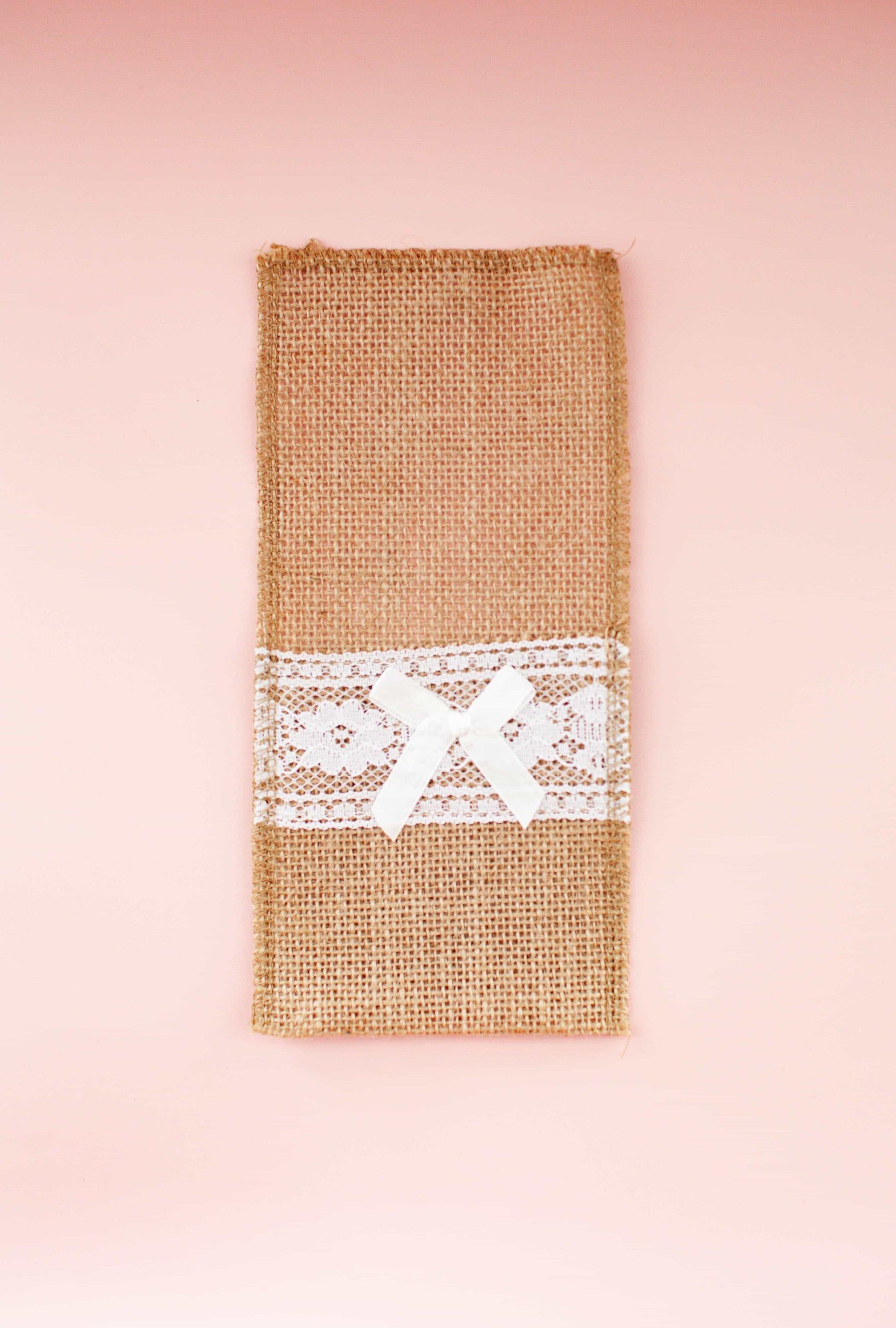 A rustic burlap wrap adorned with delicate lace and a satin bow, set against a soft pink background.