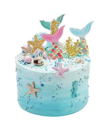 Colorful mermaid-themed cake with decorative tails, starfish, shells, and shimmering beads on a light blue frosted base.