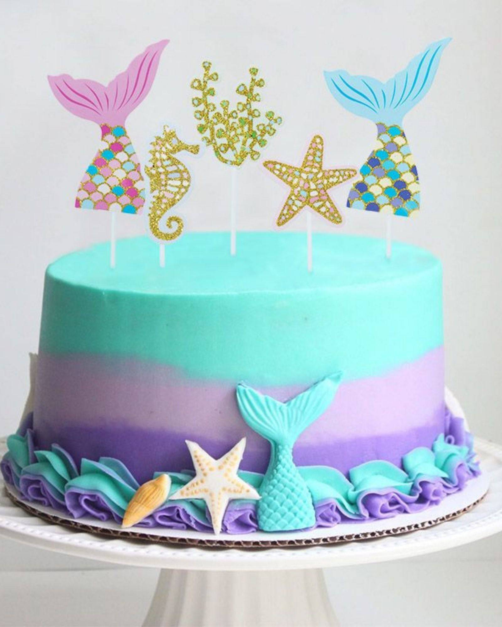 Colorful mermaid-themed decorations atop a two-tone blue and purple layered cake, featuring sea motifs and vibrant accents.