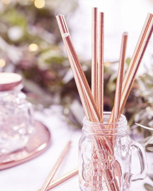 A clear glass jar filled with shiny rose gold straws, set against a softly blurred greenery background.