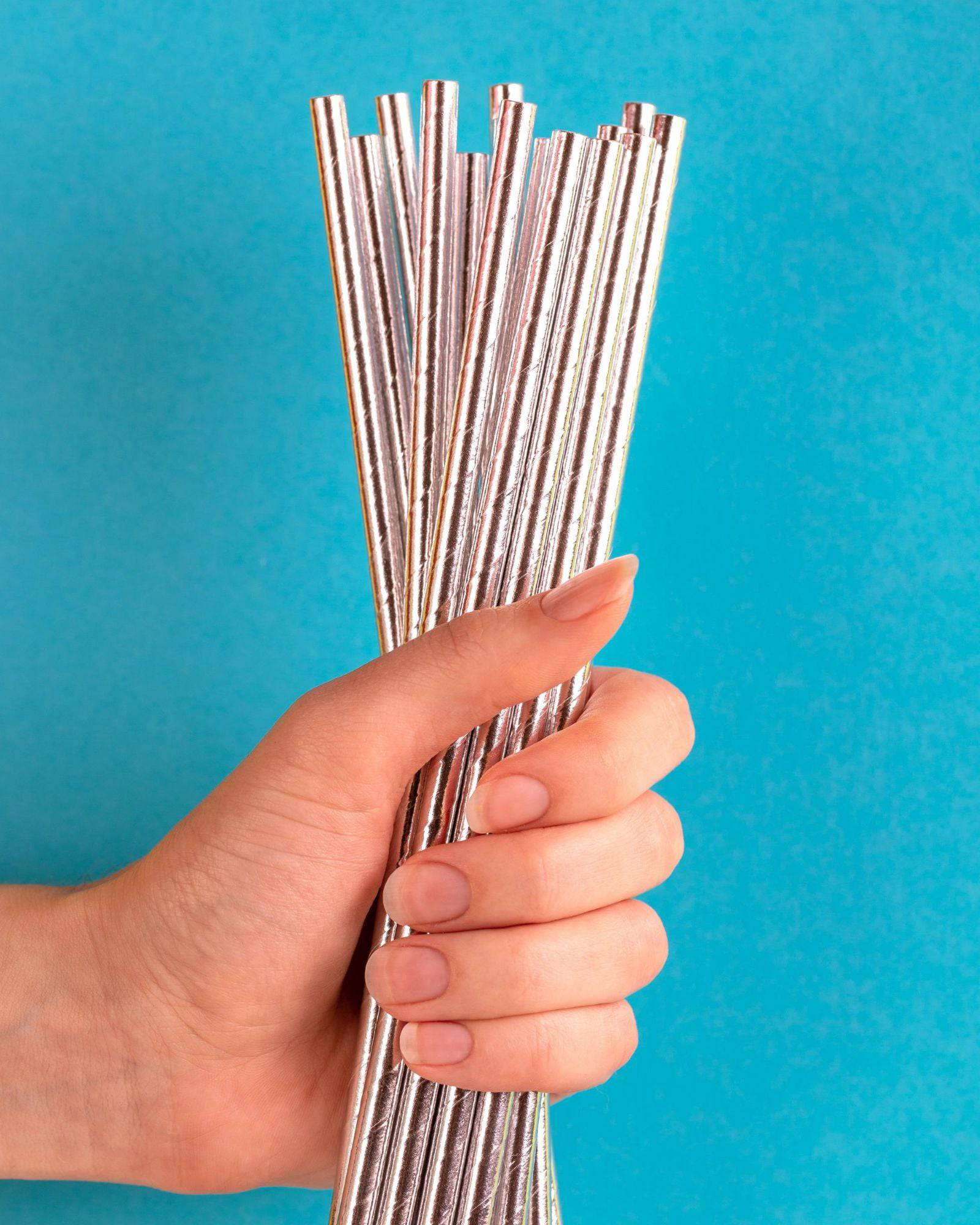 A hand holds a bundle of metallic silver straws against a bright blue background.