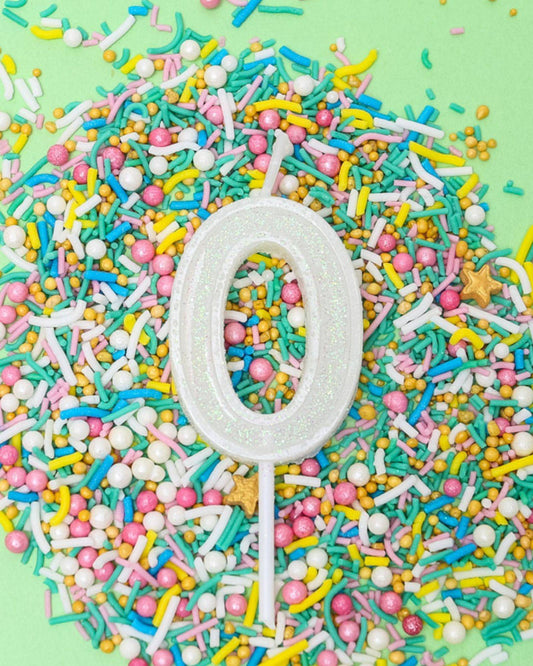 A shimmering white number zero candle surrounded by vibrant sprinkles in various colors on a pastel green background.