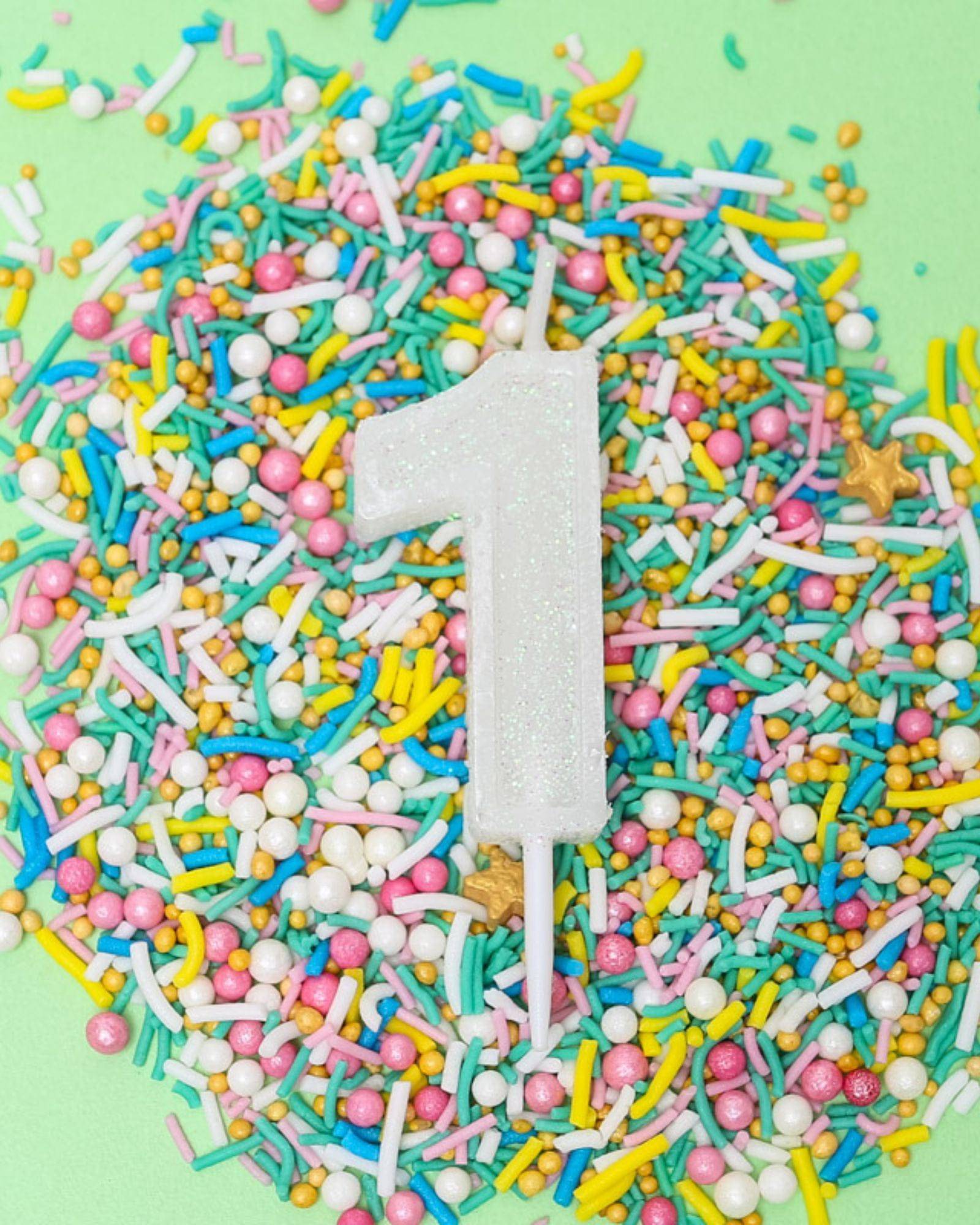 A glittery white candle shaped like the number one sits atop a colorful sprinkle mix on a pastel green background.