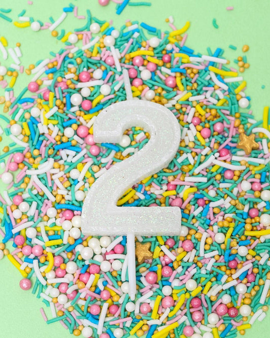 A sparkling white number two candle surrounded by colorful sprinkles on a pastel green background.