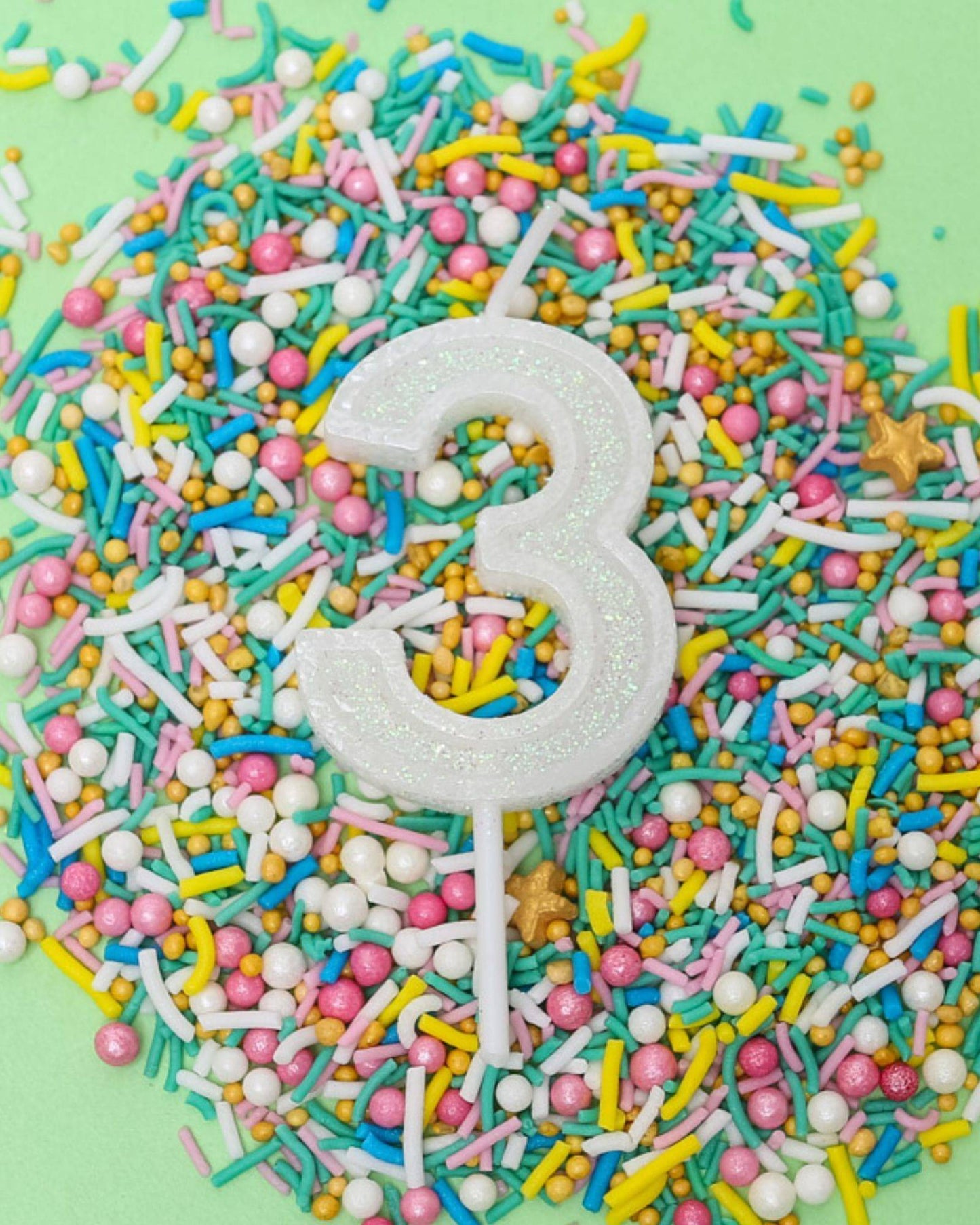 A glittery white number three candle atop a colorful array of sprinkles on a light green background.