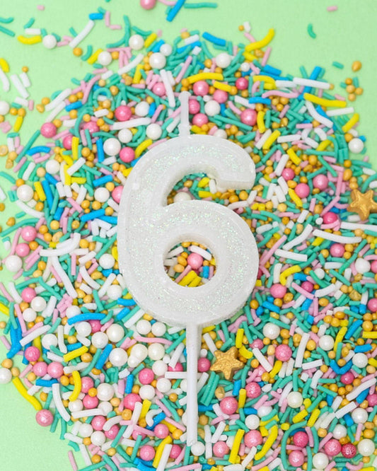 A glittery white number six candle surrounded by colorful sprinkles on a light green background.