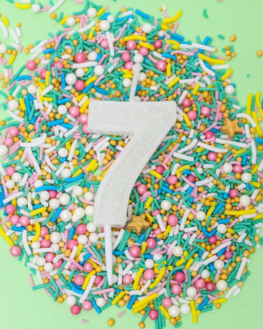 A glittery white number 7 candle surrounded by colorful sprinkles on a light green background.