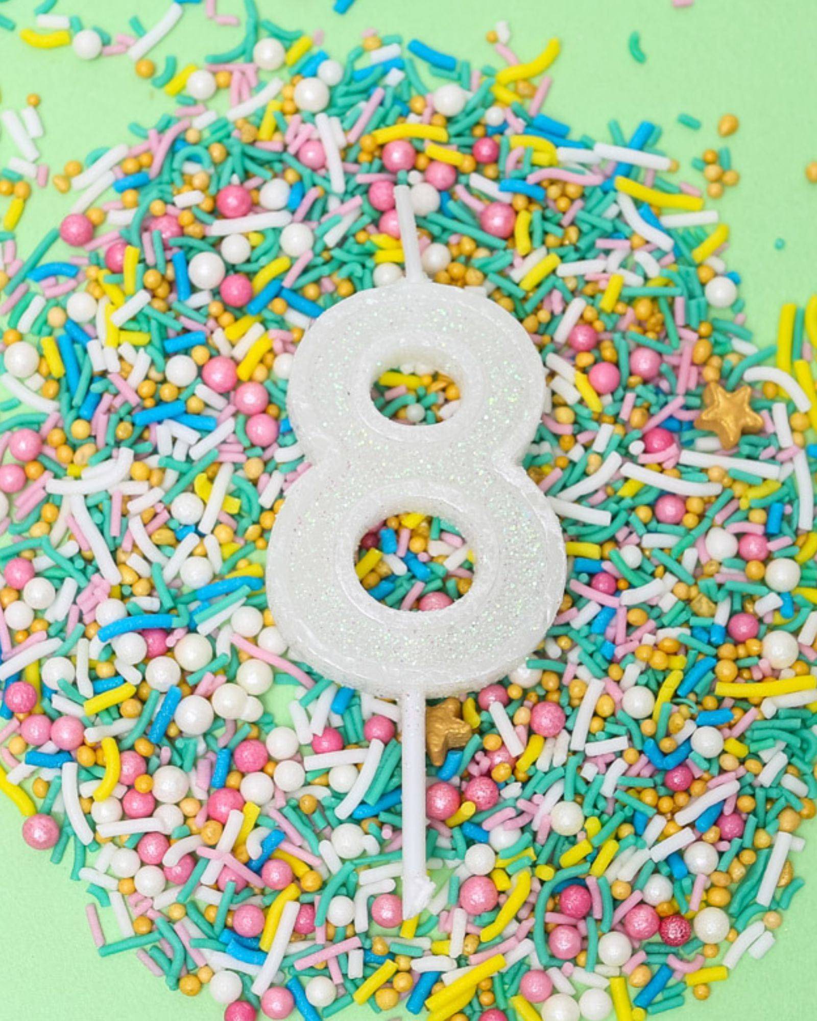 Glittery white number 8 candle surrounded by colorful sprinkles on a pastel green background.