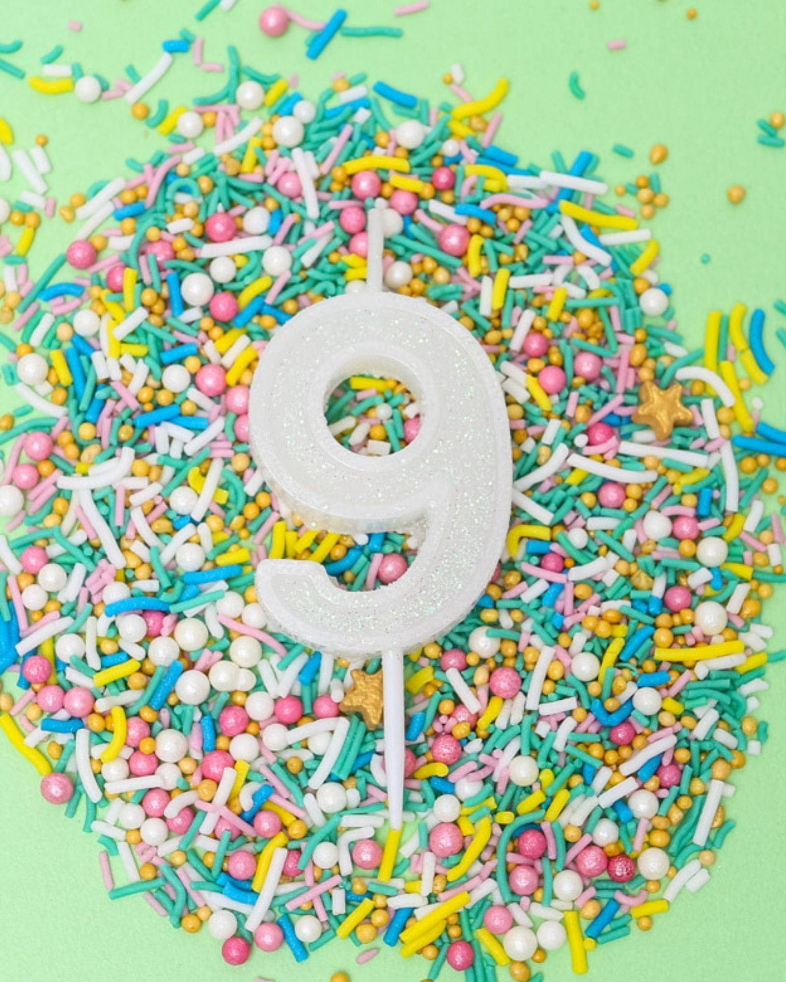 A white glitter candle shaped like the number 9, surrounded by colorful sprinkles on a light green background.