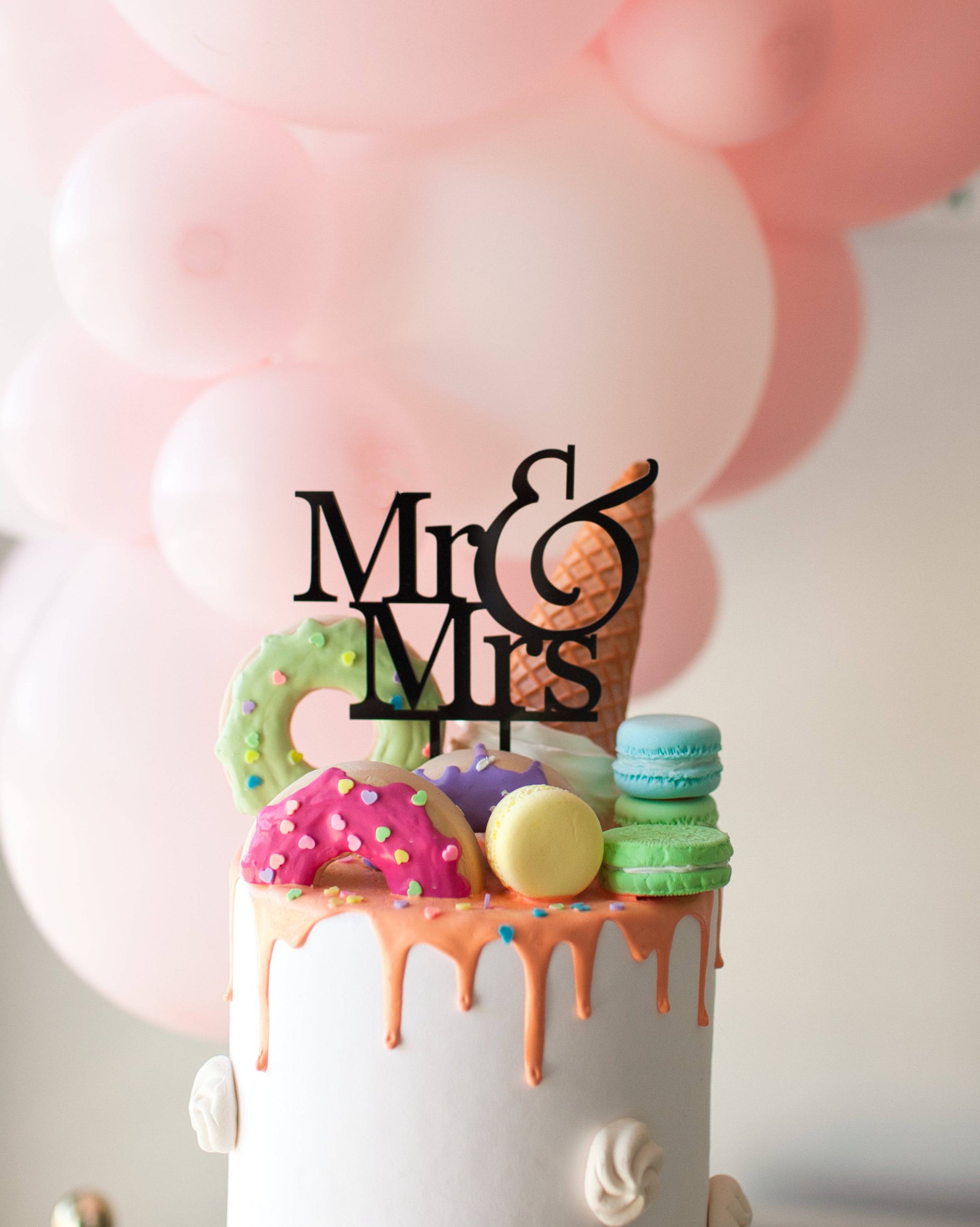 A white cake topped with colorful macaroons, donuts, and a black "Mr & Mrs" topper, against a backdrop of pink balloons.