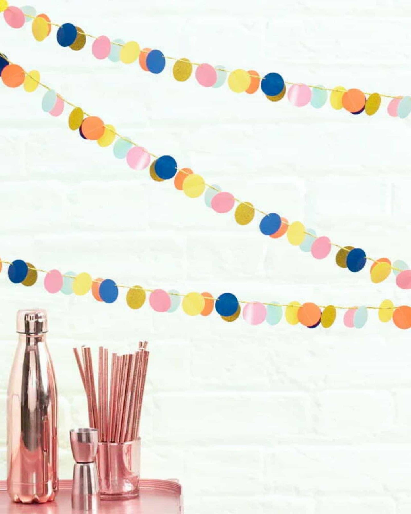 Colorful circular garland draped against a white wall, with metallic and pastel hues, beside a shiny metallic bottle and stra