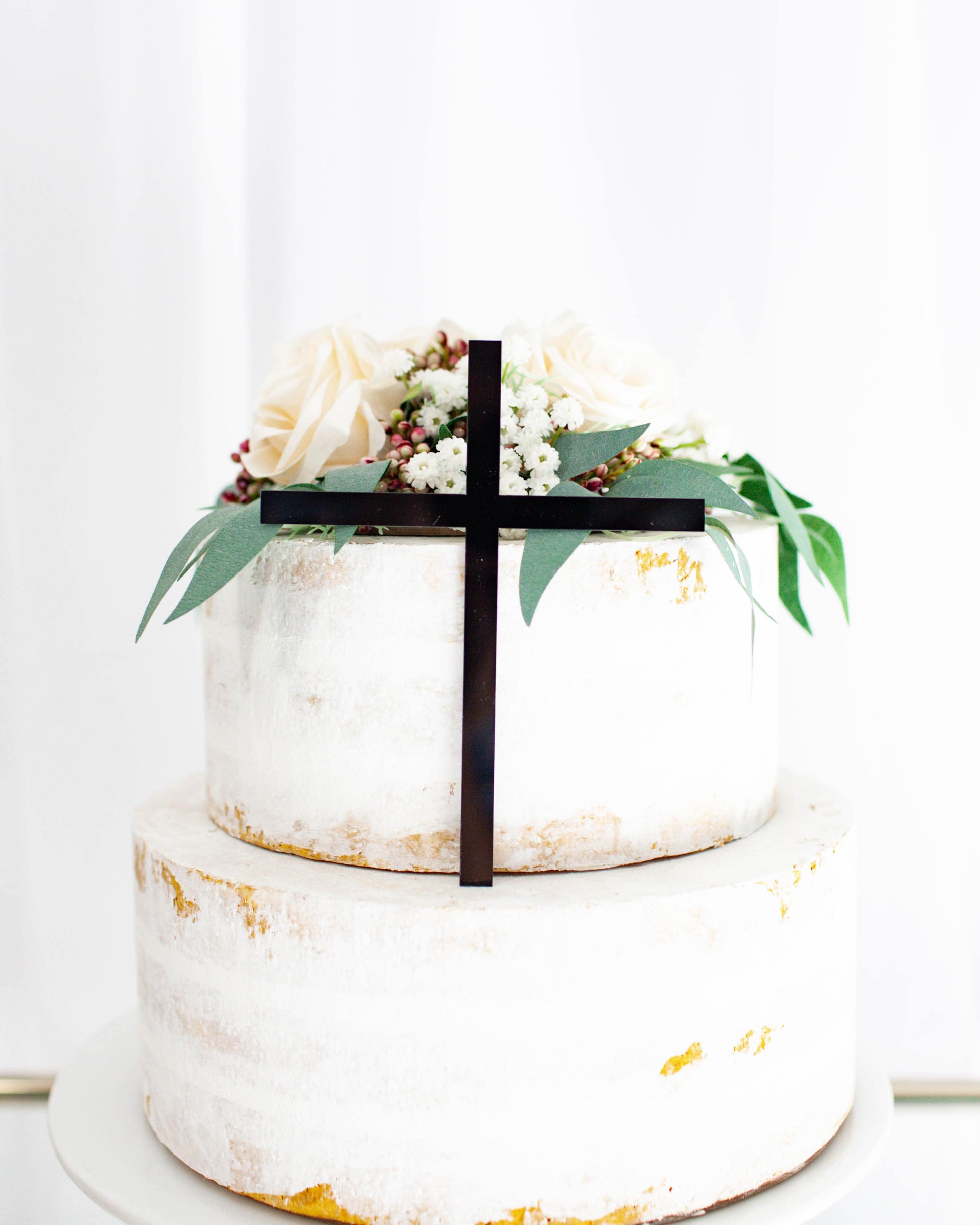 Elegant two-tiered creation adorned with soft blooms and greenery, featuring a striking black cross accent.