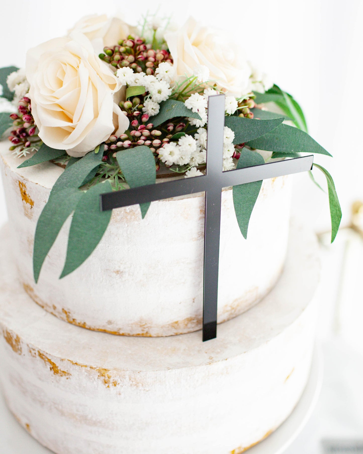 A two-tiered creation adorned with pale roses, greenery, and a sleek black cross on top, set against a light background.