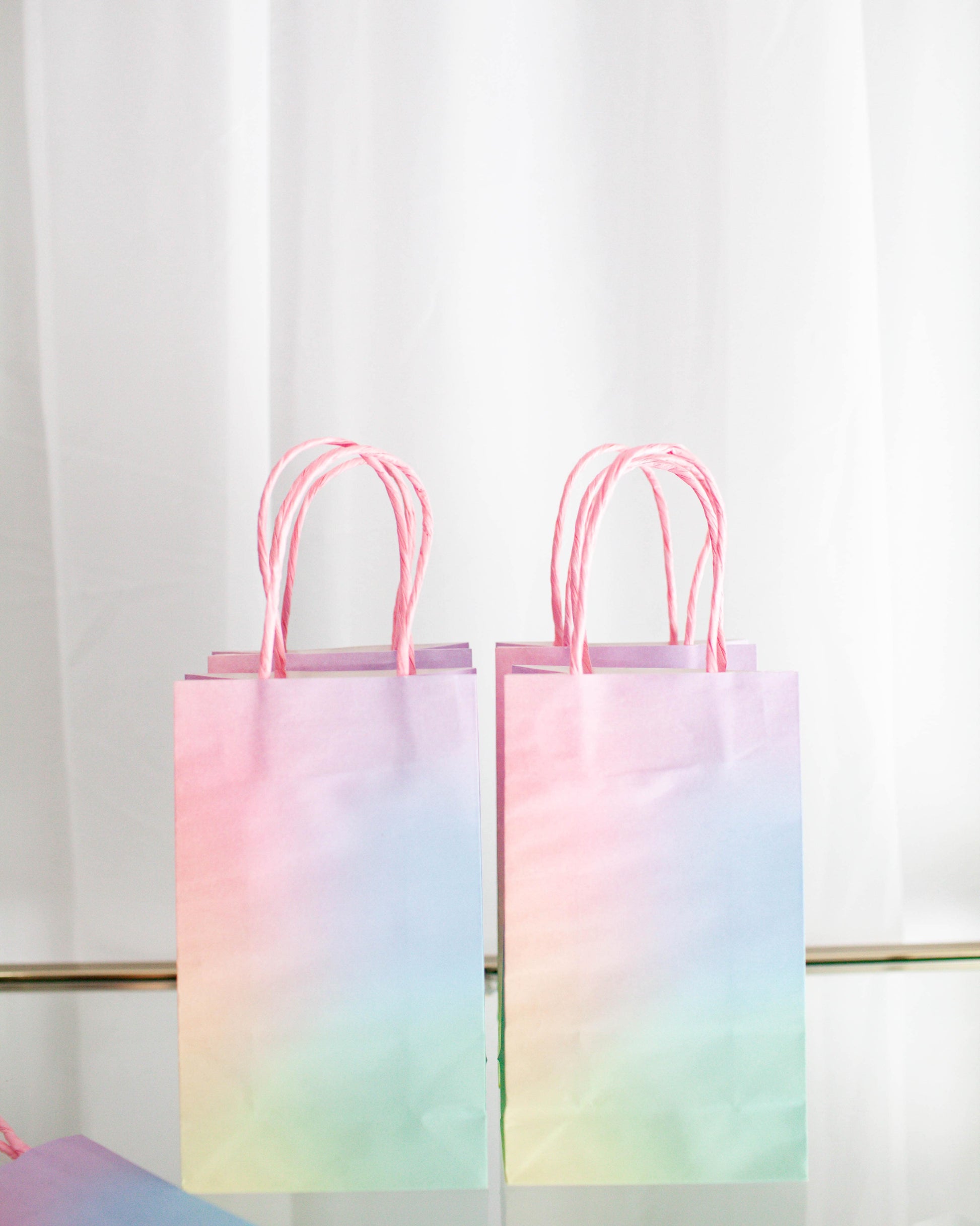 Two pastel gradient bags with pink handles against a light backdrop, showcasing soft hues of purple and green.