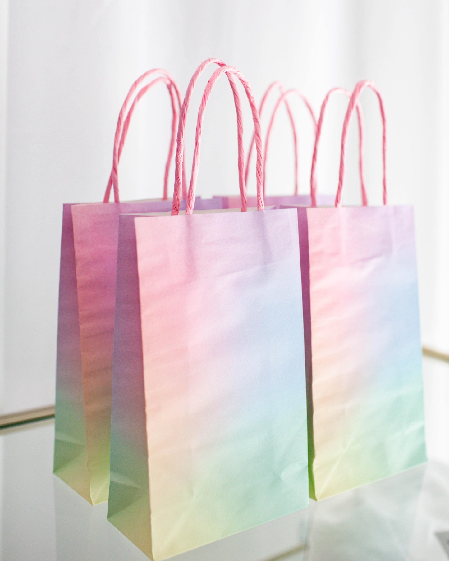 Pastel gradient bags with twisted pink handles, arranged against a bright, soft background.