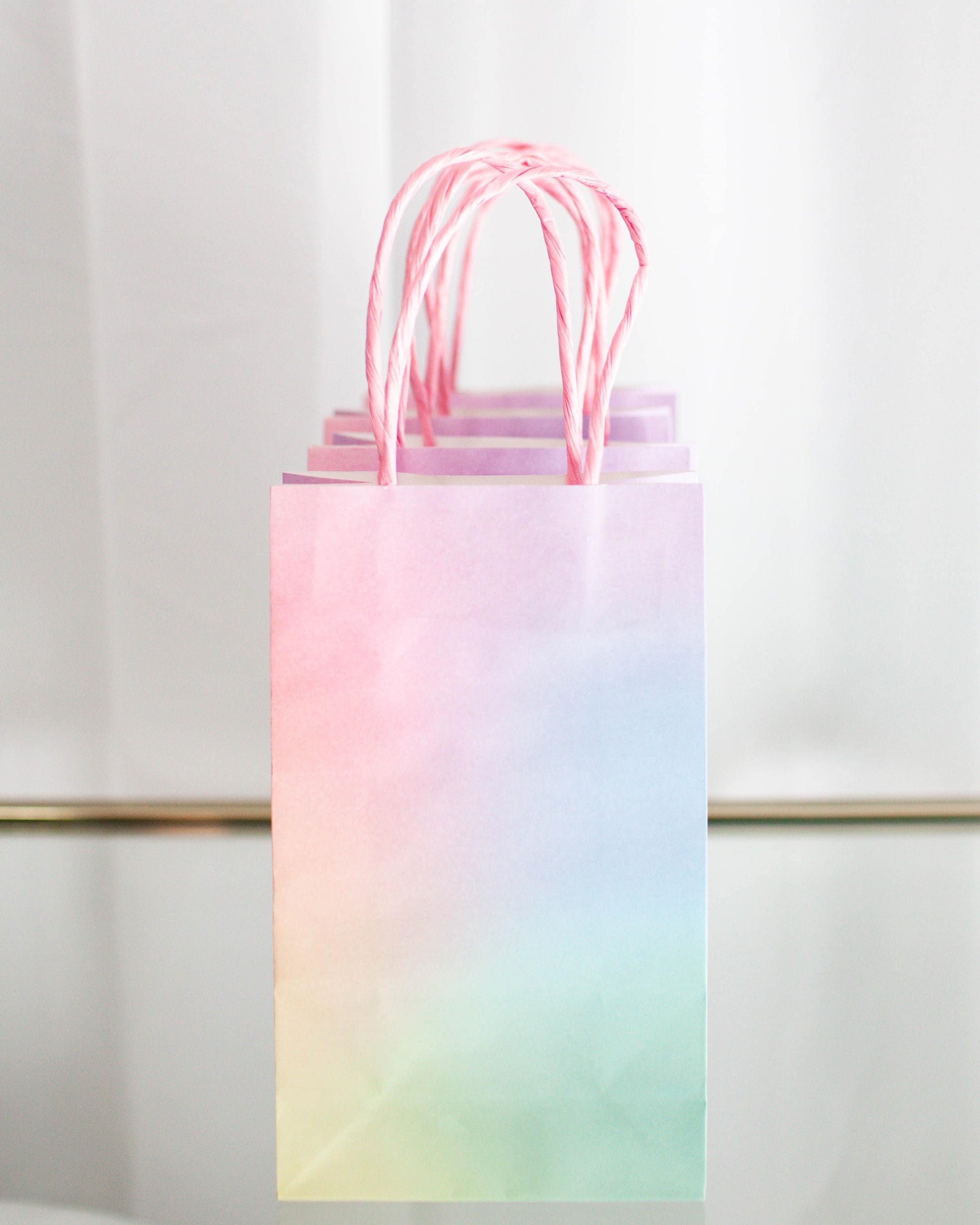 Pastel gradient bags with pink handles hanging against a soft, light backdrop.