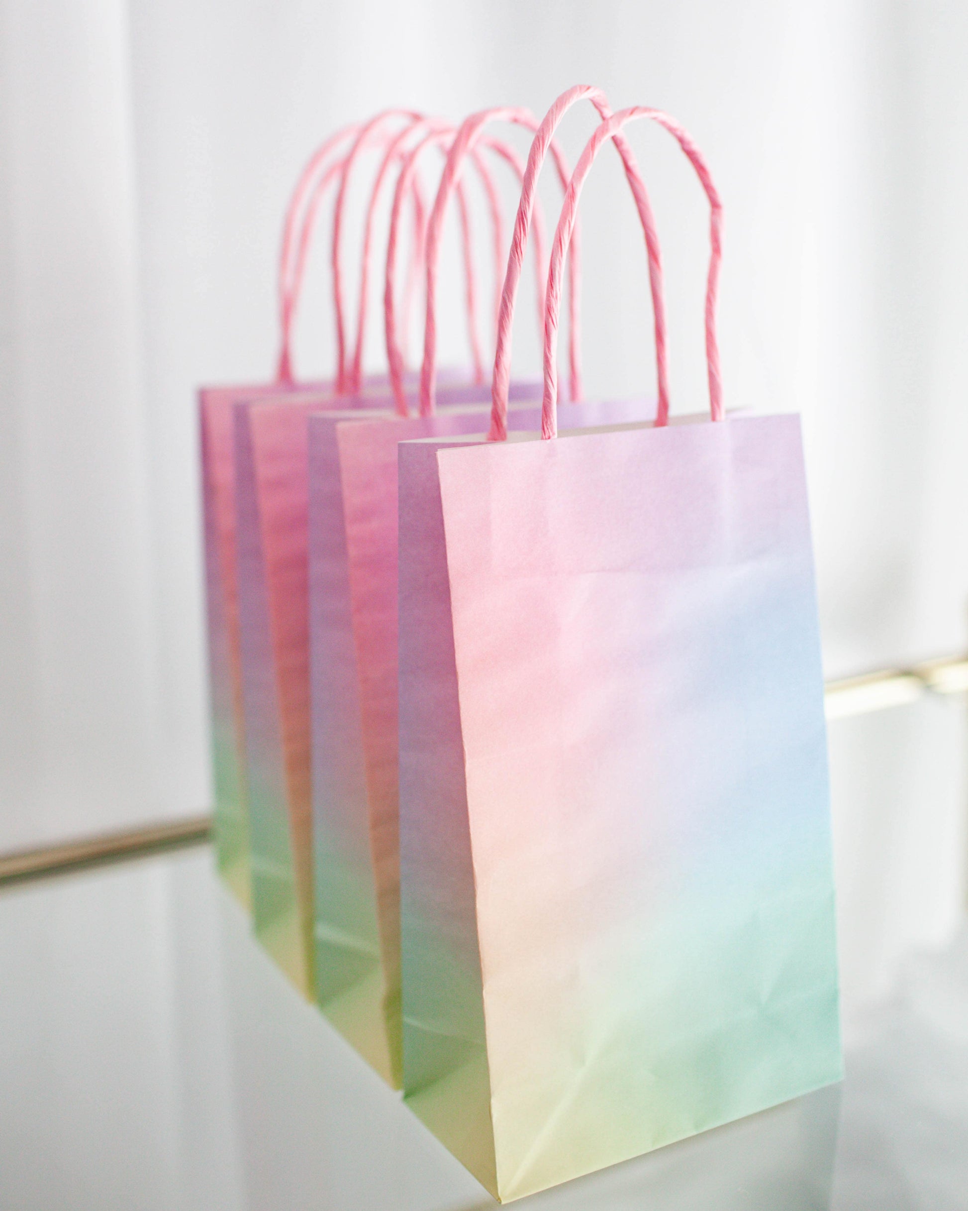 A set of pastel gradient bags with pink twisted handles against a soft, neutral background.