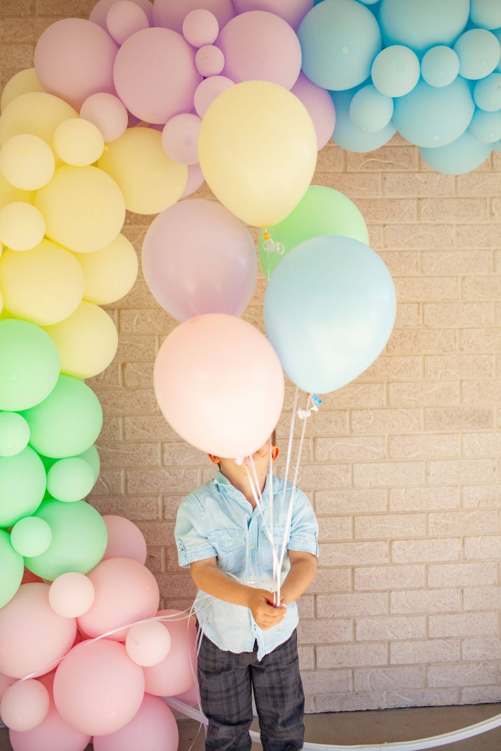 A child holds a bunch of pastel balloons in front of colorful balloon decorations against a brick wall.