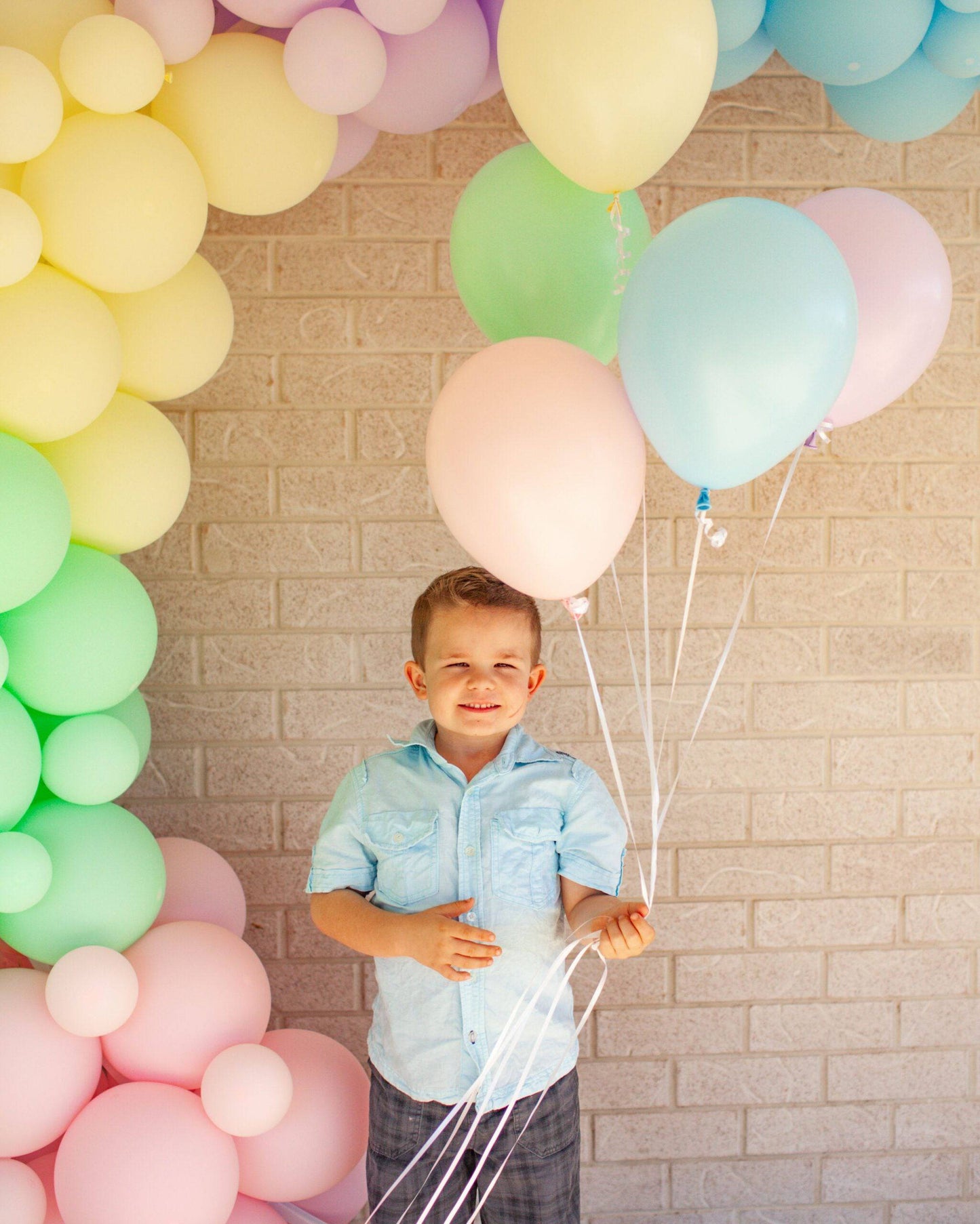 A smiling child holds colorful pastel balloons against a light brick wall, surrounded by a vibrant balloon arch.