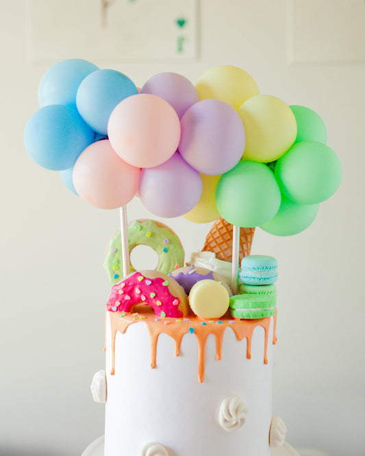 Colorful balloons atop a whimsical cake adorned with vibrant donuts, macarons, and an ice cream cone.