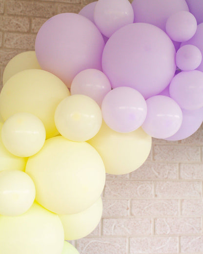 Pastel yellow and purple balloons arranged in a whimsical garland against a light brick background.