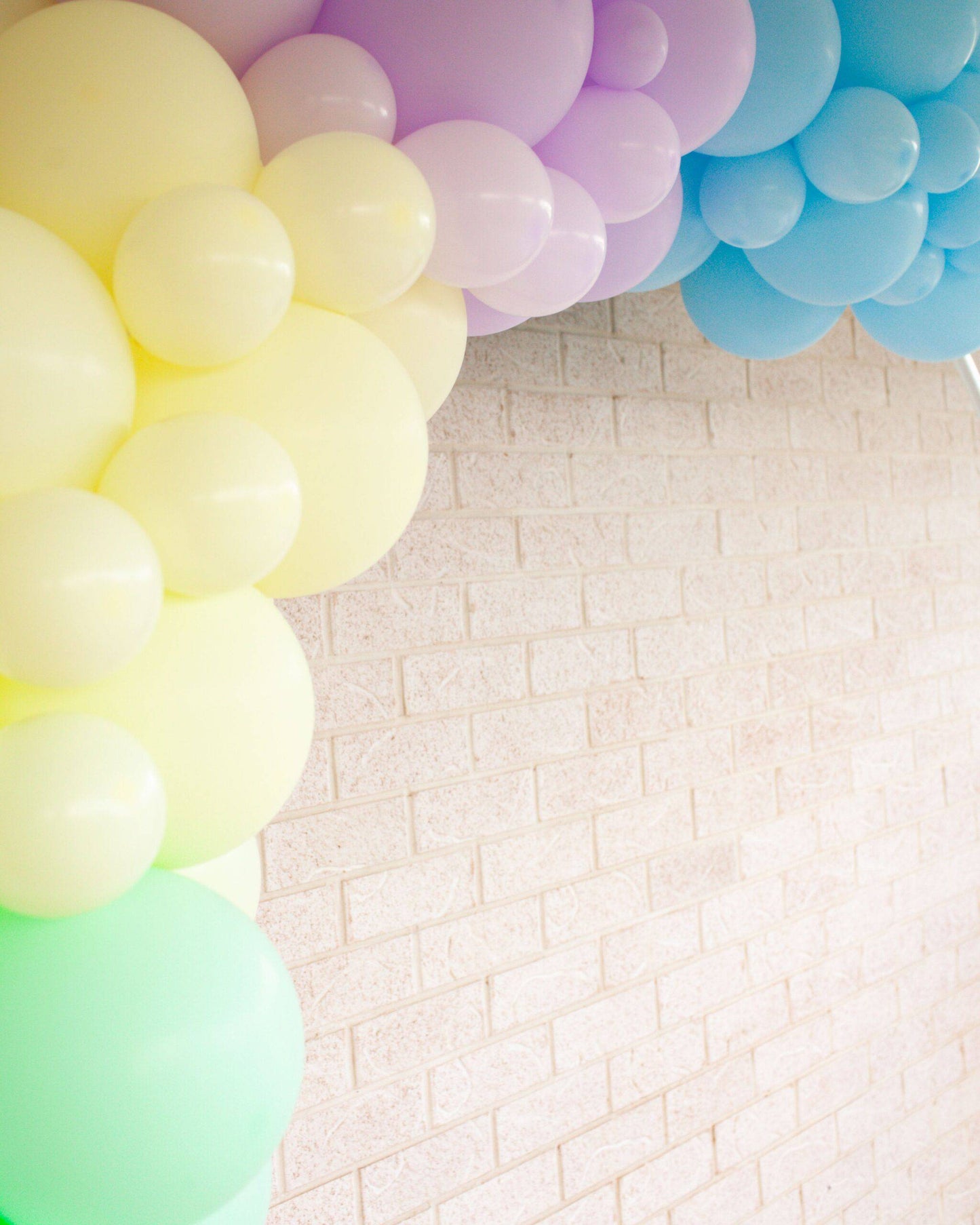 Pastel-colored balloons in yellow, lavender, and blue arranged against a textured light brick wall.