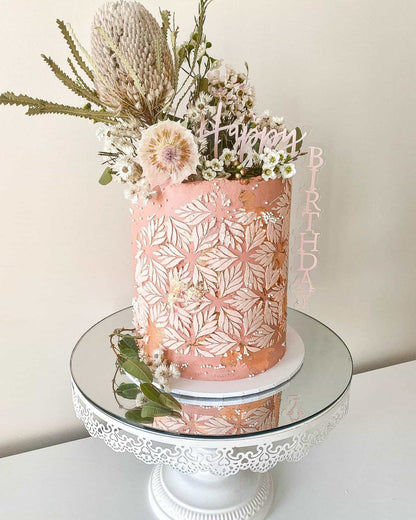 A pink cylindrical creation adorned with delicate flowers and a "Happy Birthday" topper on a reflective stand.