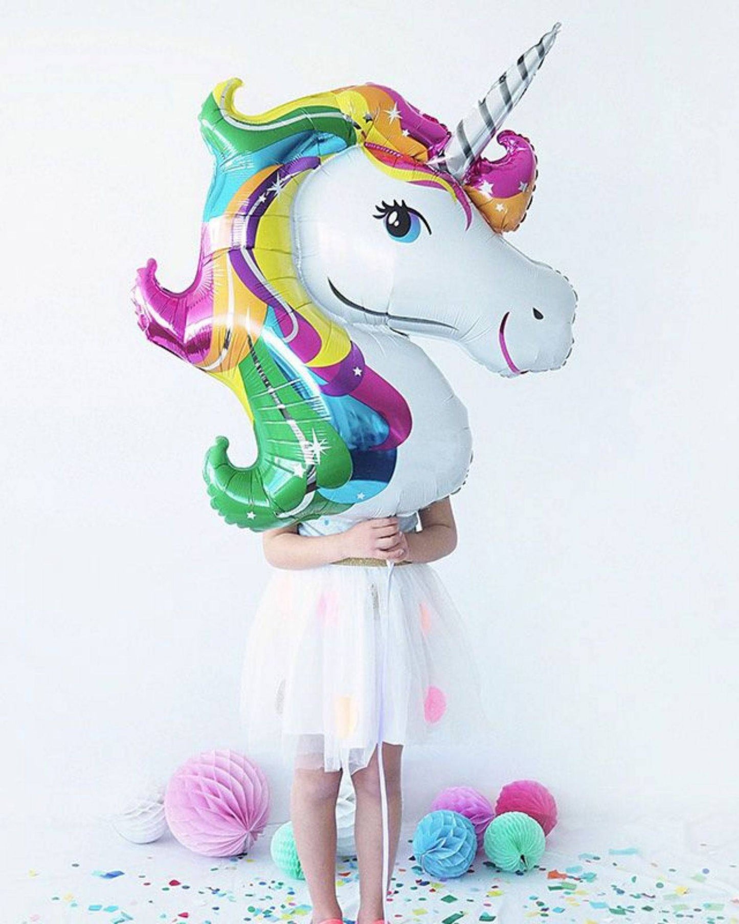 A colorful unicorn-shaped balloon held by a child in a white dress, surrounded by pastel decorations and confetti.