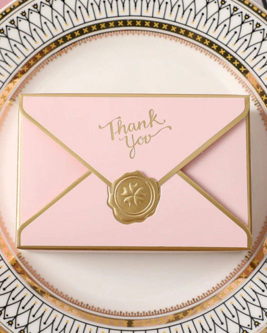 A delicate pink envelope with "Thank You" in gold, sealed with a decorative wax emblem, rests on an ornate plate.