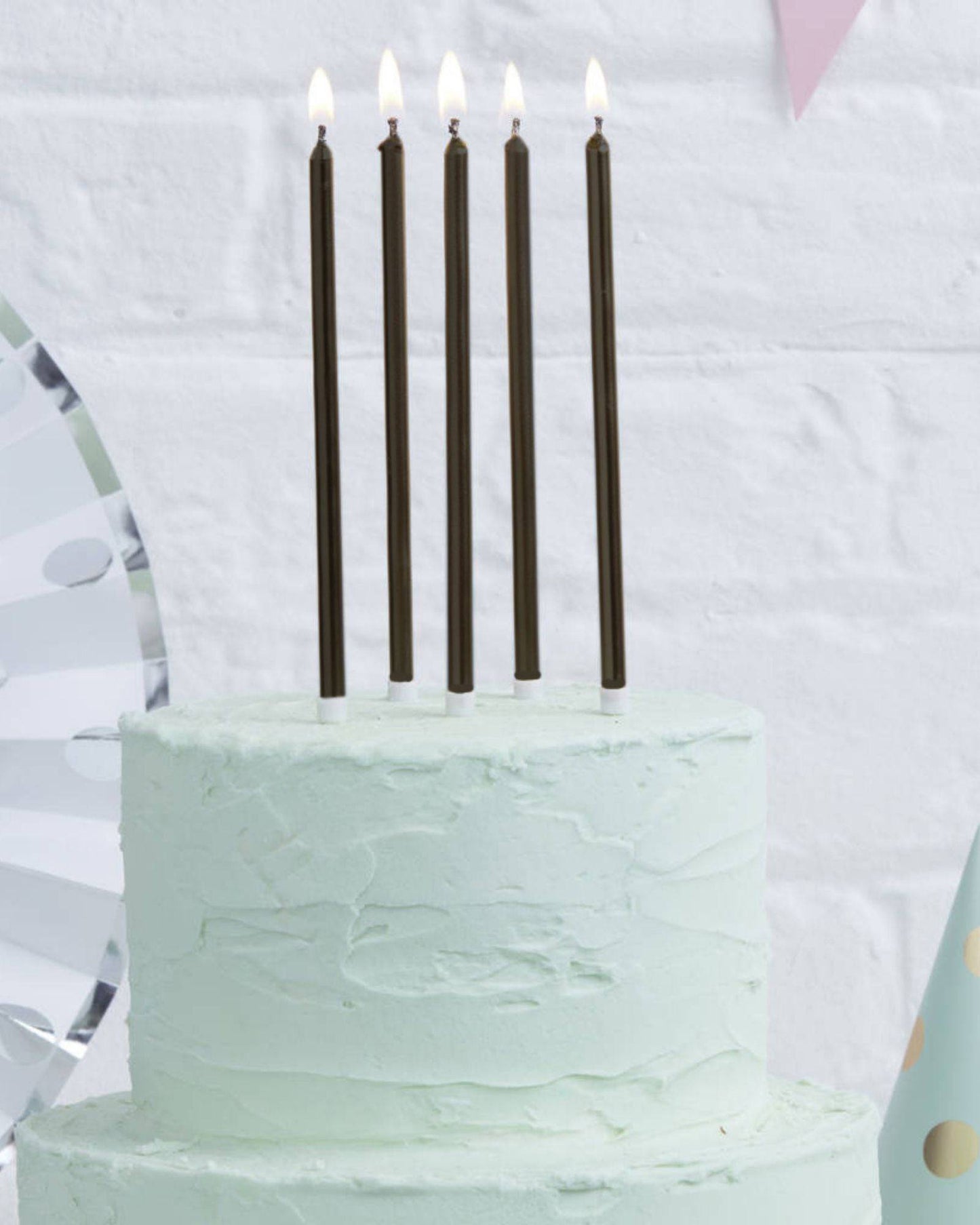 Five tall black candles with flames atop a mint green layered cake, set against a white brick background.