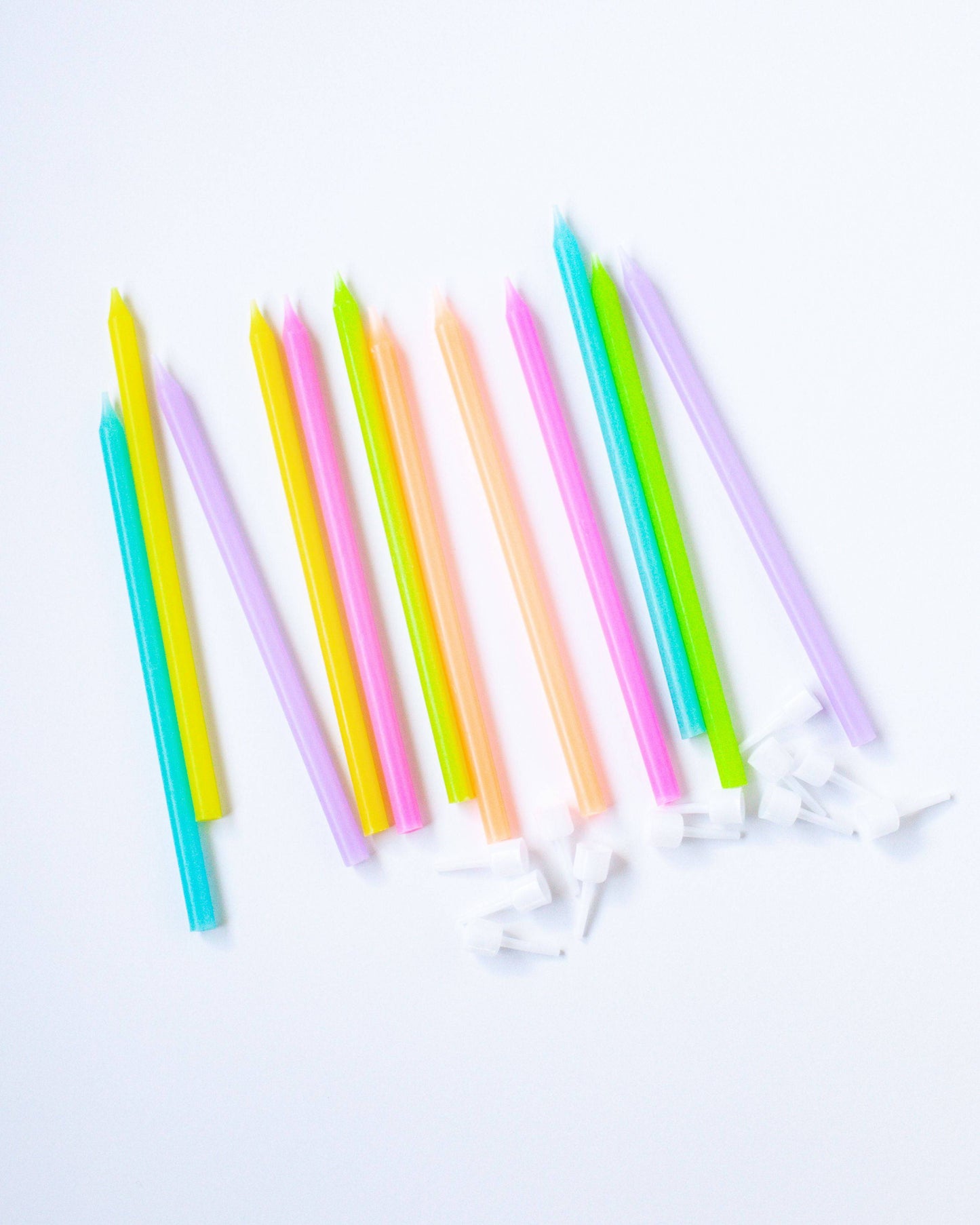 Colorful tall candles in neon hues arranged on a white background, with white plastic holders scattered nearby.