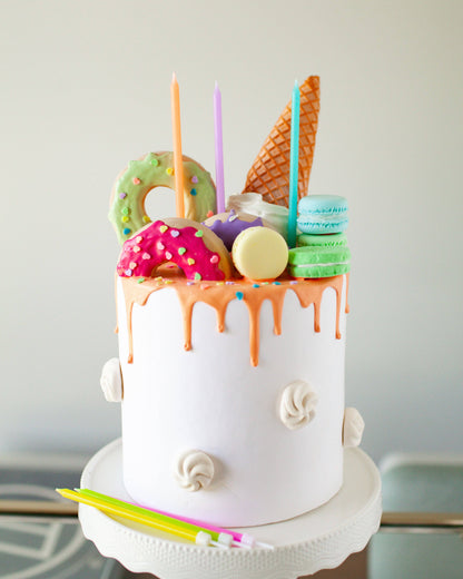 Colorful cake topped with donuts, macarons, an ice cream cone, and vibrant candles, all drizzled with orange icing.