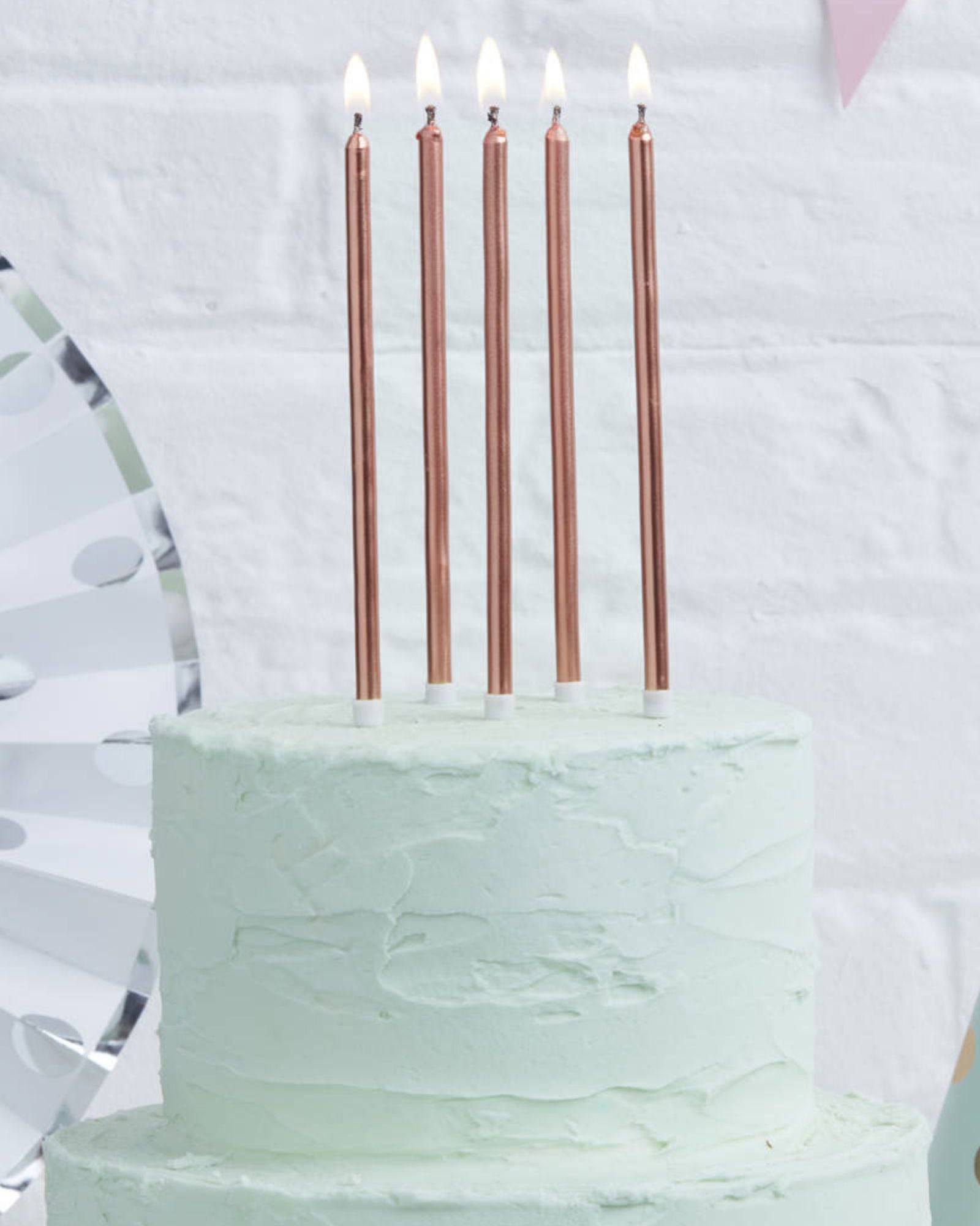 Five tall, rose gold candles with flames atop a light green frosted cake against a textured white background.