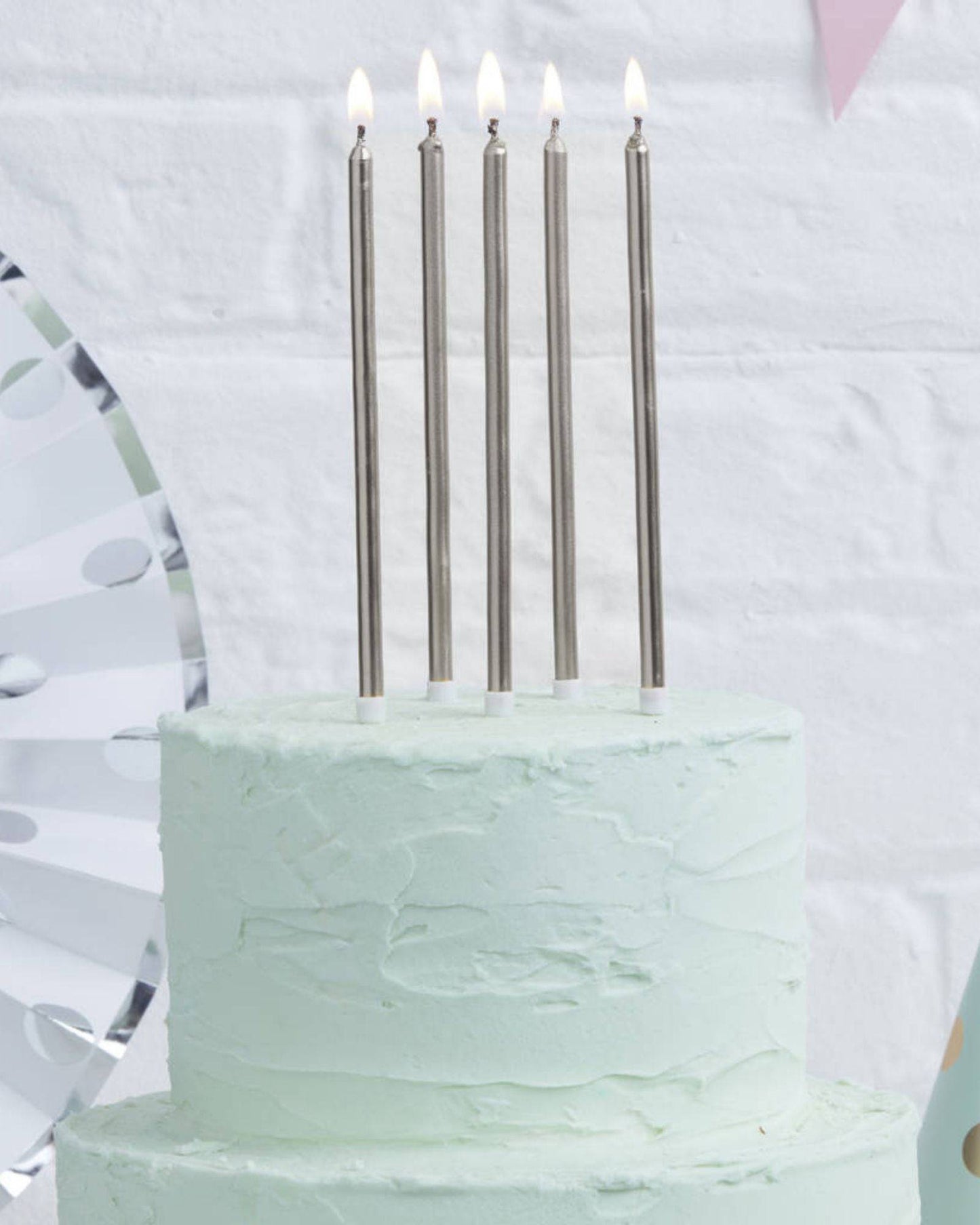 Five silver tall candles with lit tips atop a mint green frosted cake, set against a light brick background.