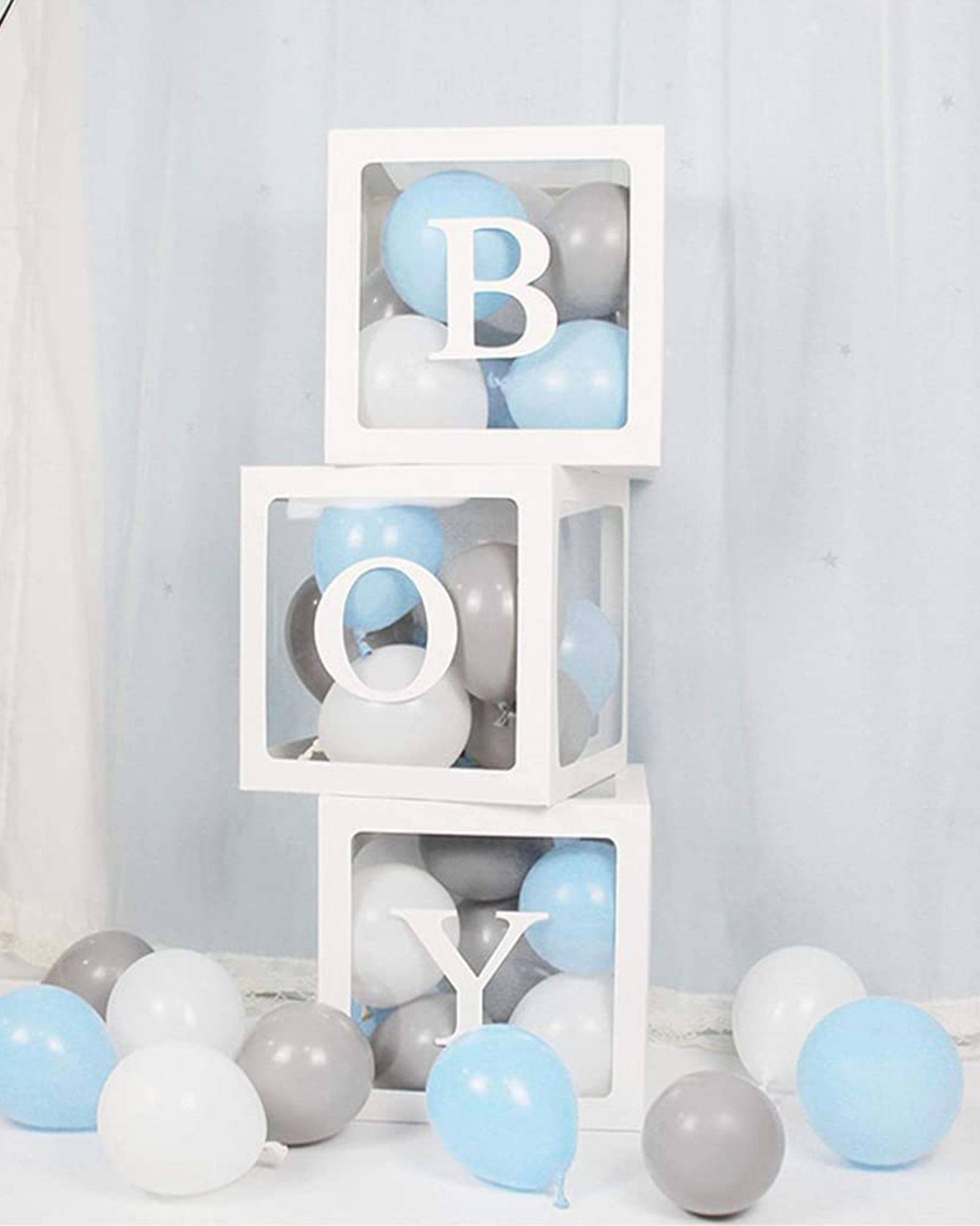 Stacked white boxes with blue, gray, and white balloons, spelling "BOY," on a soft backdrop.