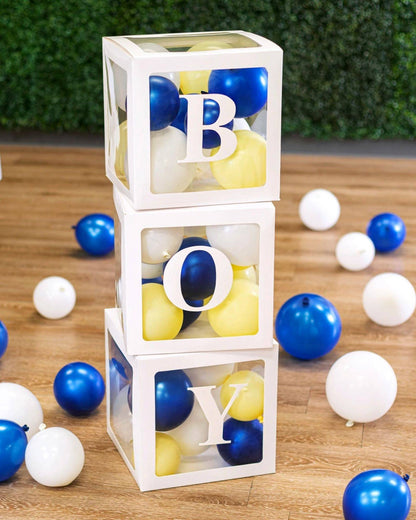 Three stacked boxes with letters "B," "O," and "Y," filled with colorful balloons, on a wooden floor.