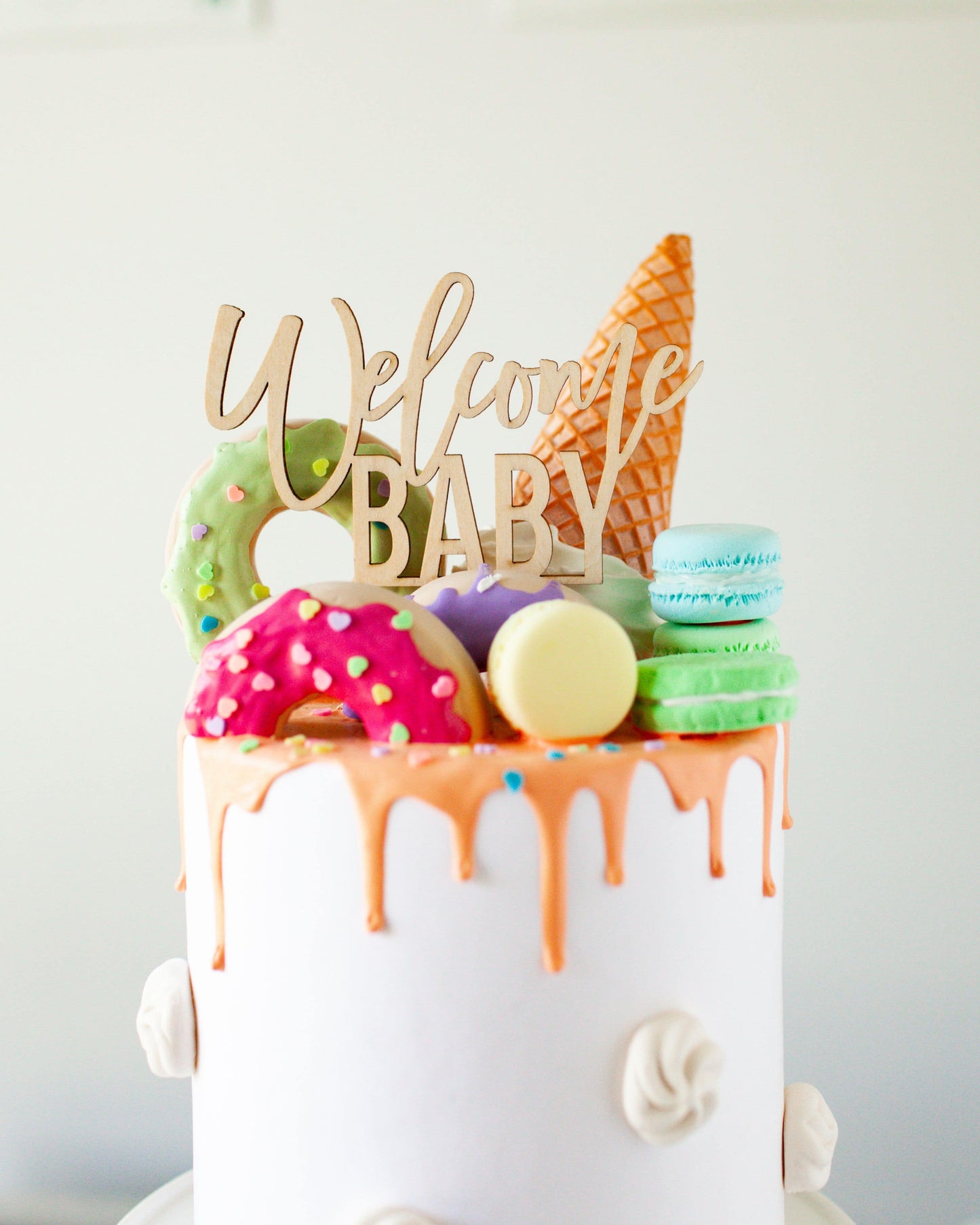 Wooden topper reads "Welcome Baby," adorned with colorful treats like donuts, macarons, and an ice cream cone.