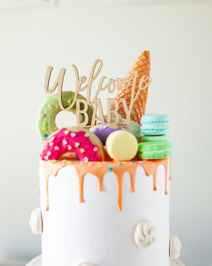 Wooden topper reads "Welcome Baby," adorned with colorful treats like donuts, macarons, and an ice cream cone.